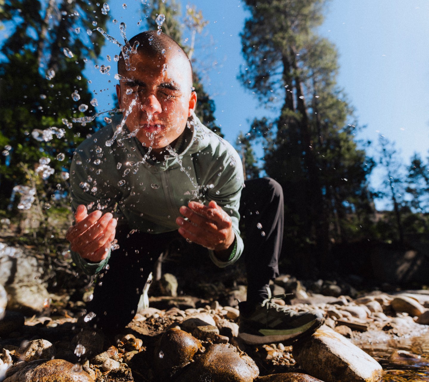 An image of a male wearing items from the new TERREX SS24 range, stooped down over a stream in a forest, splashing his face with water.