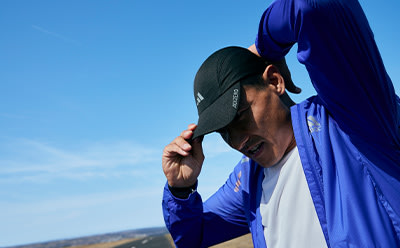 A person wearing a blue jacket and cap stands against a clear blue sky, with a distant landscape visible in the background.