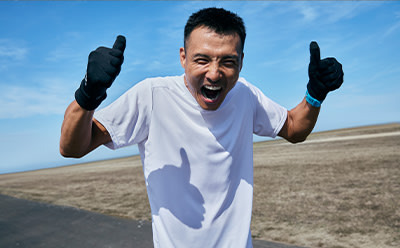 A man in a white t-shirt and black gloves is enthusiastically gesturing with his arms raised against a backdrop of a clear blue sky and a sandy, arid landscape.