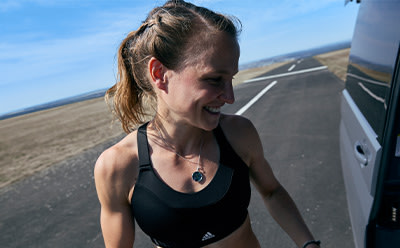 A woman with long, dark hair is smiling and looking directly at the camera, with a road and landscape visible in the background.