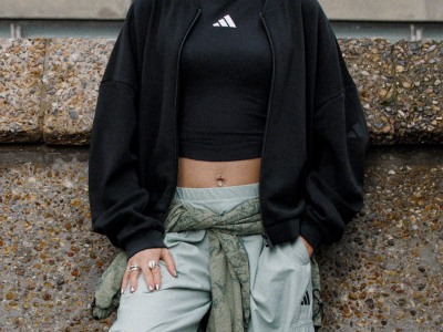 A person wearing a black jacket and crop top stands in front of a textured concrete wall, with their hands resting on their waist.