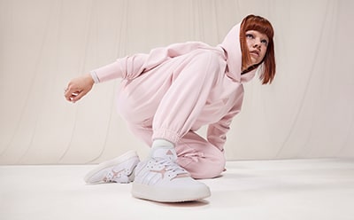 A young woman wearing a pink hooded jumpsuit and white sneakers stands in a plain, light-colored room.