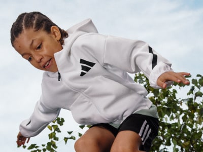 A young person wearing a white athletic jacket is crouched down and reaching towards some plants in the foreground, with a cloudy sky visible in the background.
