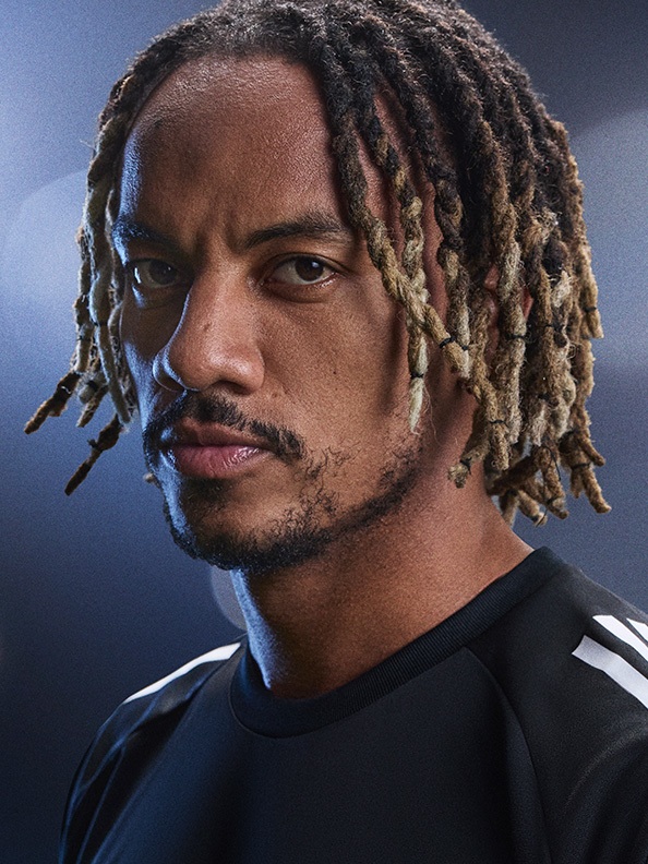 The image shows a close-up portrait of a man with dreadlocked hair against a dark blue background.