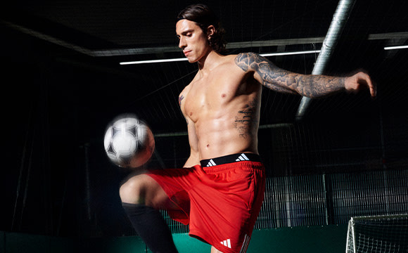 A shirtless man with tattoos is shown juggling a soccer ball in a dimly lit indoor setting.