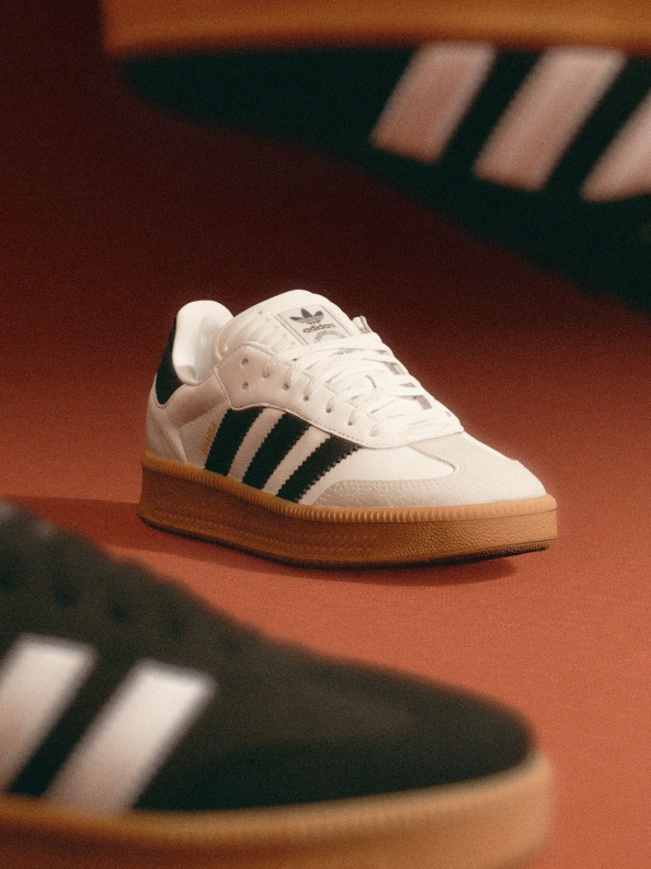 A pair of white and green Adidas sneakers resting on an orange surface, with a blurred background of what appears to be a wooden floor or table.