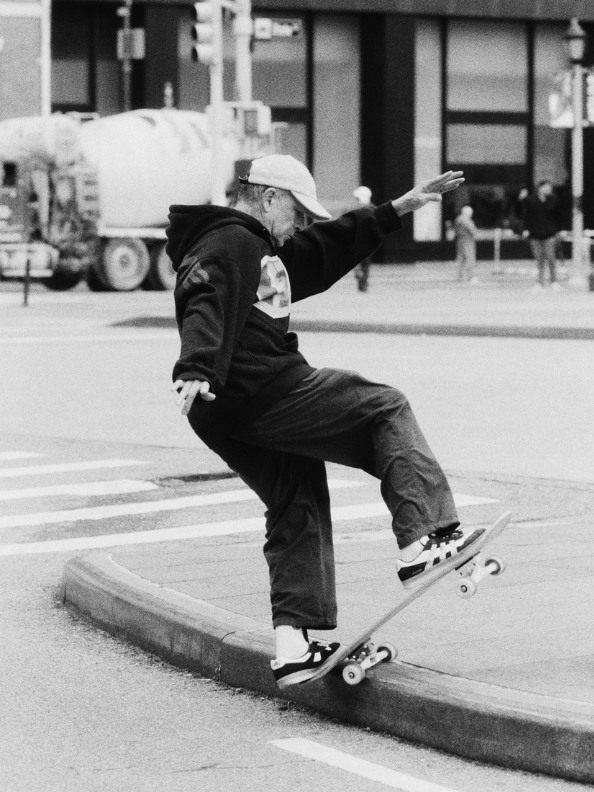 A person in dark clothing is performing a skateboarding trick on a curb in an urban setting, with buildings and a truck visible in the background.