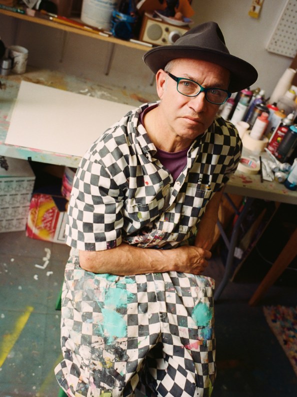 A person wearing a checkered shirt and hat is sitting in a cluttered workspace filled with various art supplies and materials.