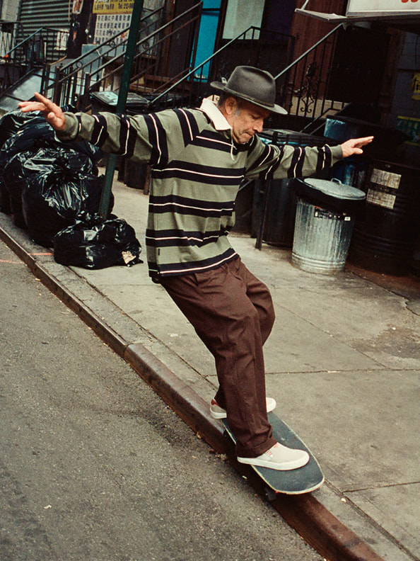 A person wearing a striped sweater and a hat is standing on a skateboard on a city street, with trash cans and other urban elements visible in the background.