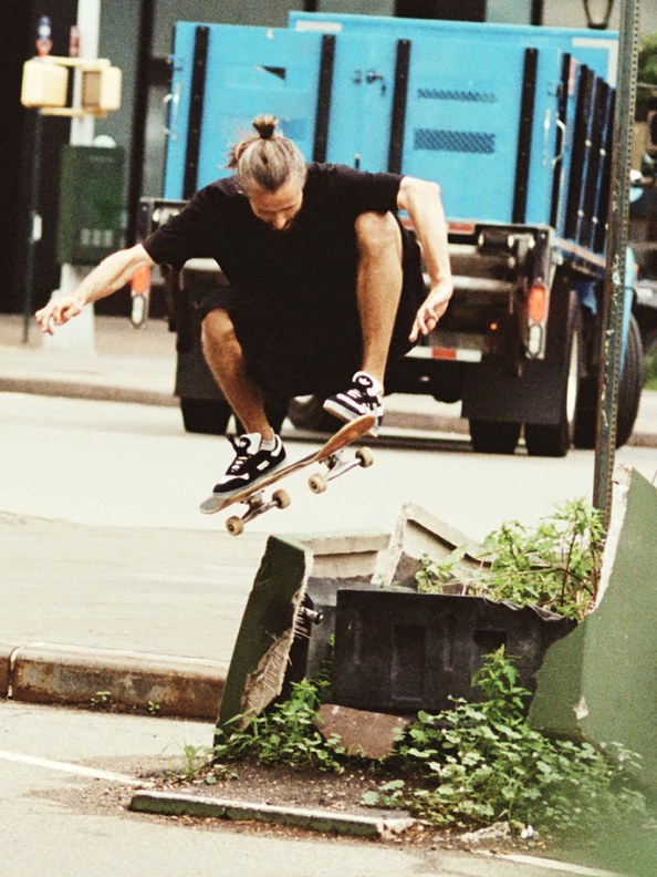 A person in black clothing is performing a skateboarding trick on a concrete ledge, with a truck and other urban elements visible in the background.