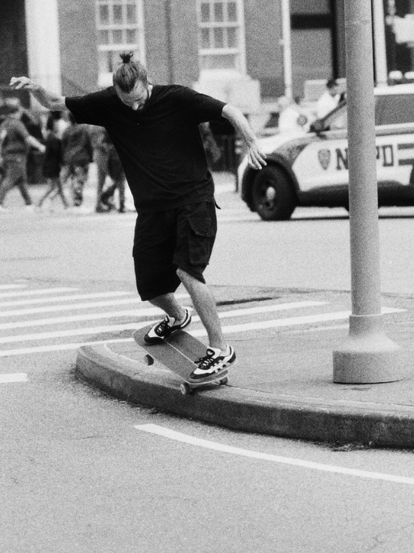 A person in dark clothing is riding a skateboard on a city street, with a police car visible in the background.