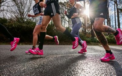 The image shows several people running on a paved path in a wooded area, wearing bright pink athletic shoes.