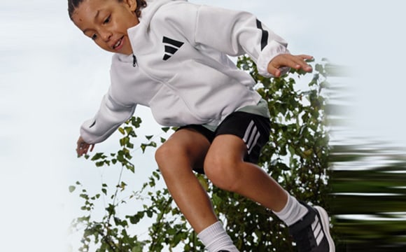 A young person in a white athletic jacket and black shorts is running through a blurred, green environment.