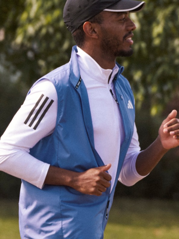 A man with dark hair running in nature wears a white adidas long sleeve shirt, a blue vest and a black adidas cap.