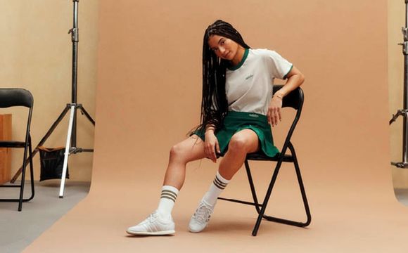 A person with long dark hair, wearing a white shirt and green shorts, sitting on a chair in a studio setting with a plain beige background.