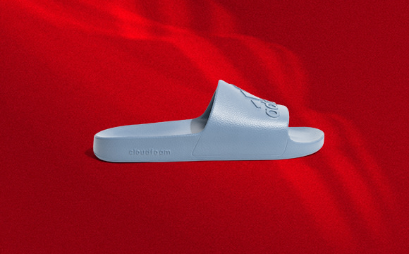 A white slide sandal with the Adidas logo sits on a vibrant red background.