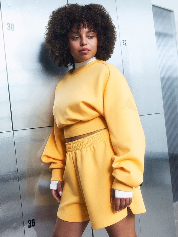 Una mujer joven de pelo oscuro rizado con una sudadera y un pantalón corto amarillos se coloca de pie frente a una pared o una ventana de cristal, con una expresión neutra en la cara.