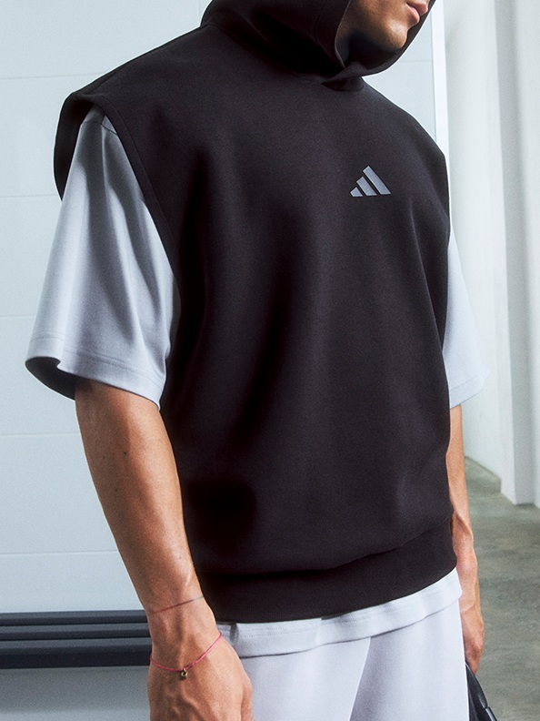 Před bílou zdí stojí člověk v černé mikině adidas.