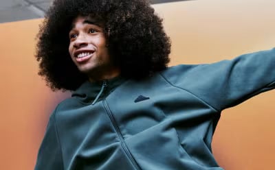 A person with curly dark hair wearing a green jacket appears to be smiling and gesturing in a brightly colored setting.