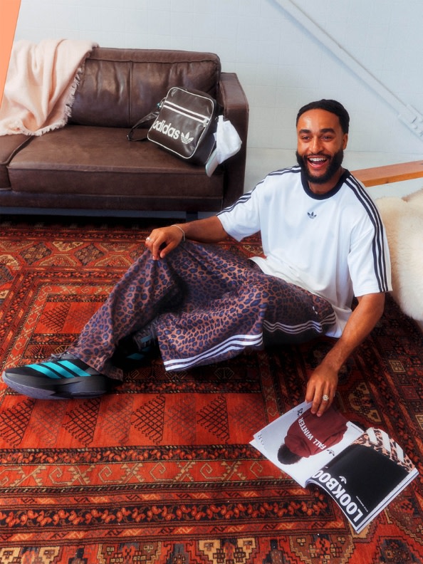 Ein lächelnder Mann sitzt auf einem farbenfrohen gemusterten Teppich in einem Raum mit einem braunen Ledersofa und anderen Möbeln.