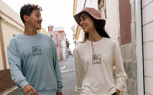 Two people, a man and a woman, are standing in an urban setting, wearing casual clothing with a similar logo design.