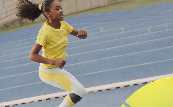 A young girl in a yellow shirt and white pants is running on a blue track surrounded by a fence.