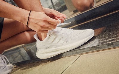 The image shows a person's hands tying the laces of a pair of white sneakers on a tiled floor.