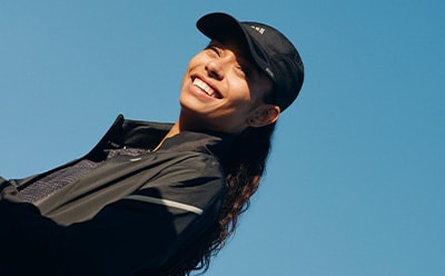 A person wearing a black jacket and cap is smiling against a clear blue sky.