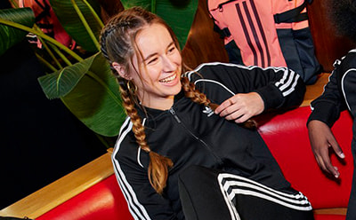 A young woman with braided hair is smiling and wearing a black athletic jacket with white stripes in a colorful, vibrant setting.