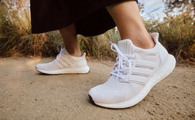 The image shows a person's feet wearing white athletic shoes standing on a dirt path surrounded by tall grass.