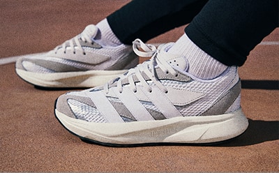 The image shows a pair of white and gray athletic shoes with thick soles, worn by a person standing on a wooden floor.