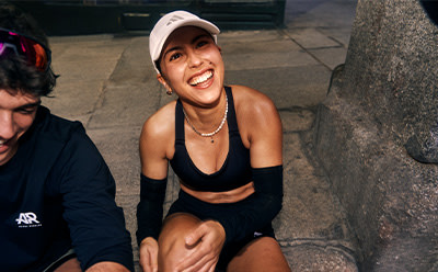A smiling young woman in athletic clothing sits on a stone wall, with a city street and buildings visible in the background.