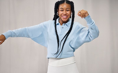 A smiling young woman with braided hair wearing a light blue sweater and white pants, standing against a plain background.