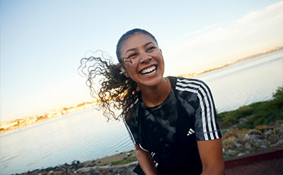 A smiling young woman with curly hair stands in front of a scenic coastal landscape at sunset.