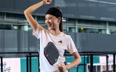 A young woman in a white athletic top is smiling and raising her arms in a celebratory gesture, with a modern glass and steel building in the background.