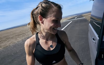 A woman with long, dark hair is smiling and looking directly at the camera, with a road and landscape visible in the background.