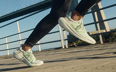 The image shows a person's legs and feet wearing white and green sneakers standing on a wooden deck with a body of water and buildings visible in the background.