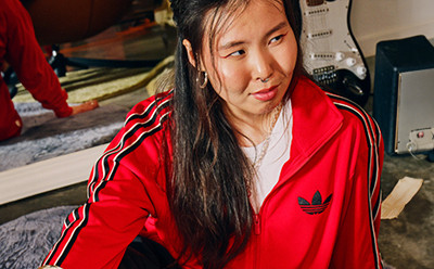 The image shows a young woman wearing a bright red Adidas jacket with white stripes, standing in what appears to be a cluttered room or studio setting.