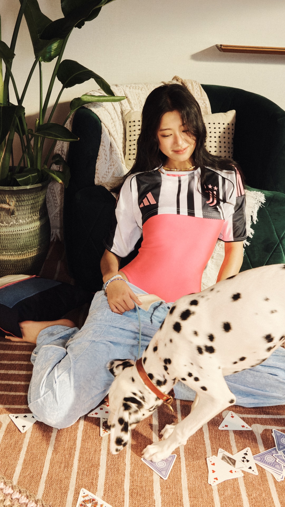 A young woman sits on the floor surrounded by various objects, including a potted plant, a pencil, and scattered papers, creating a cozy and creative atmosphere.