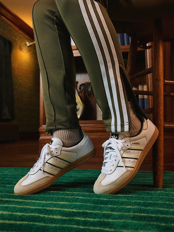 The image shows a person's legs and feet wearing white sneakers with three stripes, standing on a green carpet in what appears to be a room with wooden furniture and shelves in the background.