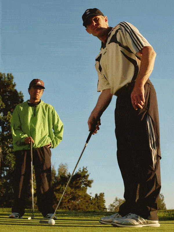 Two older men, one wearing a green jacket and the other wearing a beige jacket, are standing on a golf course with trees and a clear sky in the background.