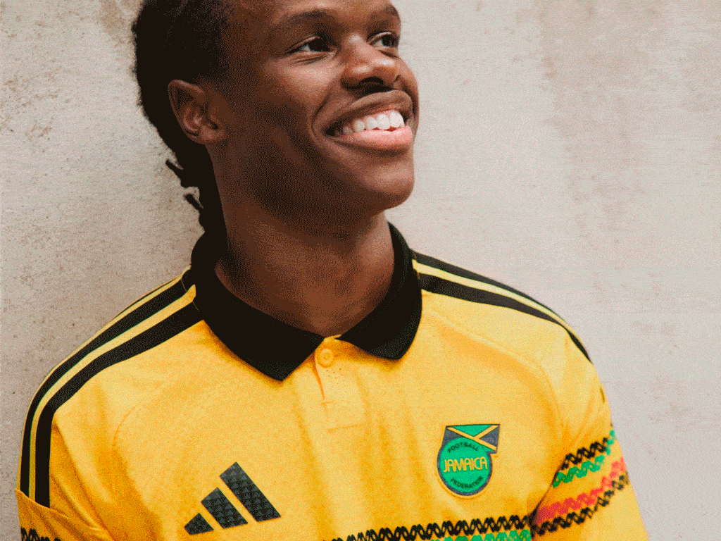 A young man wearing a yellow and black sports jersey with the Jamaican flag emblem, smiling brightly against a plain background.