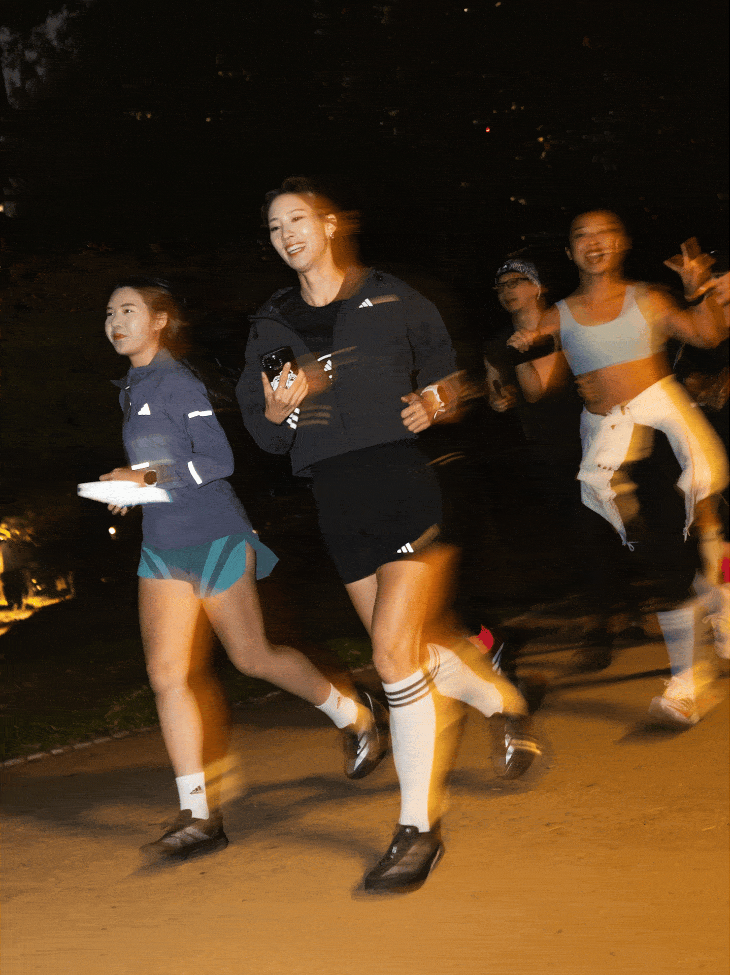 The image shows a group of people, likely young adults, engaged in a nighttime outdoor activity such as running or jogging, with the background appearing to be a dimly lit park or outdoor setting.