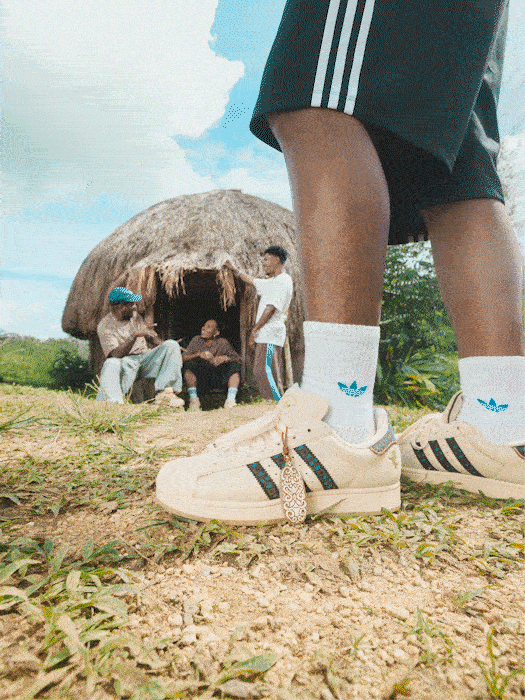 A thatched hut stands in a grassy field, with a person wearing Adidas shoes and socks visible in the foreground.