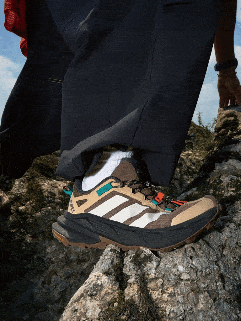 The image shows a person's lower body wearing dark pants and hiking boots standing on a rocky, mountainous terrain with a cloudy sky in the background.
