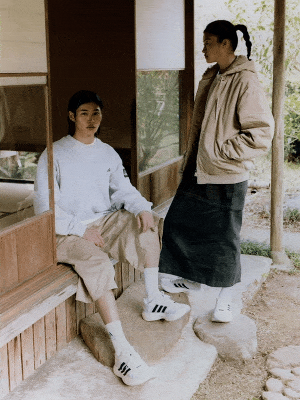 Two individuals, a man and a woman, are seated on a wooden bench in what appears to be a traditional Japanese-style building with a view of the outdoors through a window.