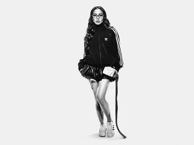 A woman with long dark hair stands in a black and white image, wearing a black tracksuit top and shorts, and holding a cane.