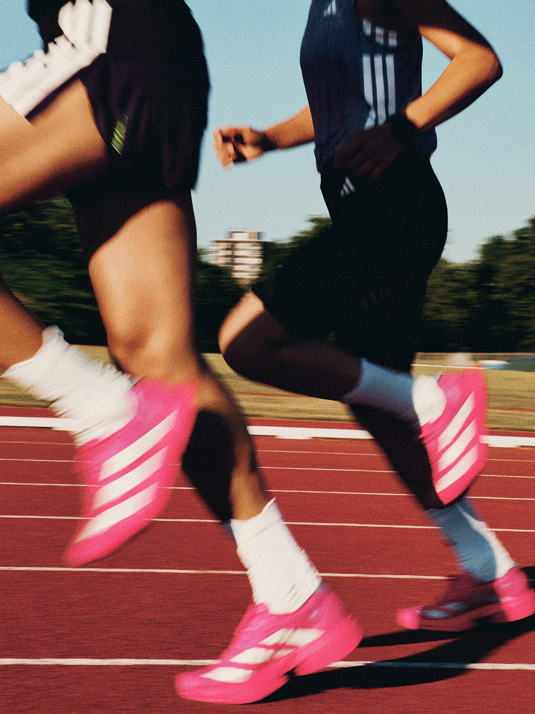 The image depicts a person wearing black athletic clothing and bright pink sneakers running on a red track surrounded by a blurred outdoor environment.