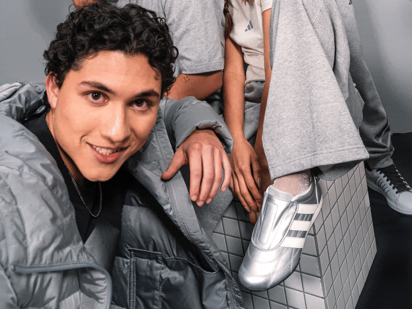 A smiling individual with curly dark hair wearing a gray jacket and silver shoes, standing in front of a gray background.