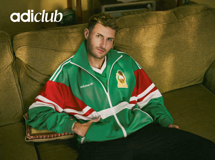 A man wearing a green and red sports jacket is sitting on a beige couch in what appears to be a living room or lounge area.